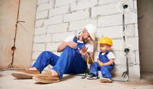 Male Builder In Work Overalls Showing Pliers To Little Boy In Apartment Under Renovation. Man And Kid In Safety Helmets Sitting By Brick Wall In Empty Building. Home Renovation And Parenting Concept.