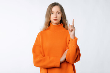 Wall Mural - Serious woman pointing finger up, showing smth upsetting, looking annoyed. Lovely female model wears trendy knitwear orange sweater standing against neutral studio background