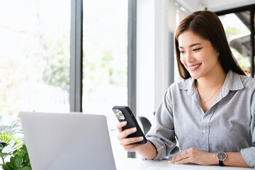 Wall Mural - Cheerful woman entrepreneur sitting beside a window and smiling. Businesswoman working in office with a mobile phone in hand.
