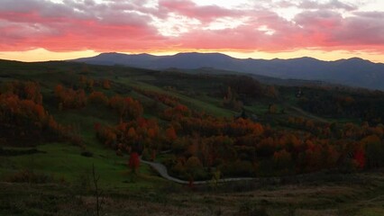 Canvas Print - Landscape of valley of green hills with colorful trees under red cloudy sky at sunset