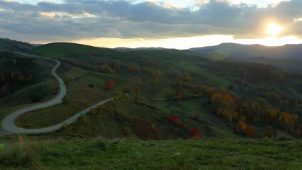 Wall Mural - Drone landscape view of a curvy road on green valley hills with hazy sky at sunset