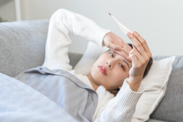 Wall Mural - Sick, influenza asian young woman, girl have a fever, hand holding thermometer, touching forehead for check measure body temperature, illness while lying rest on sofa at home. Health care concept.