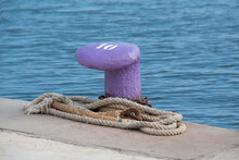 Bollard Or Mooring Bollard Installed In A Port, Purple With The Number 10, With An Old Rope And Rusty Orange Chains At Its Feet With A Beautiful Blue Sea In The Background