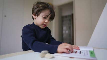 Wall Mural - Small boy reading book sitting at desk in bedroom. Curious child studying by himself turning page and looking at text and images