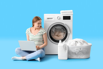 Canvas Print - Pretty young woman with modern laptop sitting near washing machine on blue background