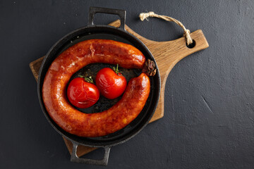 Wall Mural - Fried beef sausage tied with a ring and baked tomatoes in a black cast iron pan on a wooden cutting board on a black stone table. Top view with copy space