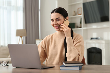 Canvas Print - Happy woman talking on smartphone while working with laptop at desk in room