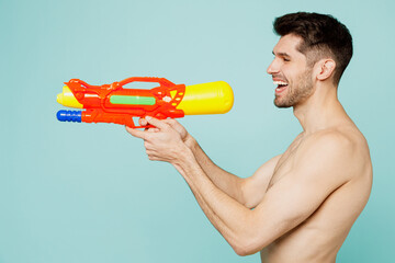 Side view young man wear green shorts swimsuit relax near hotel pool hold in hand play shoot from water gun play game isolated on plain light blue background. Summer vacation sea rest sun tan concept.