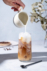 Iced coffee latte in a tall glass with a hand pouring milk from a milk jug on a blue background with coffee beans, branch and morning shadow. Summer refreshment concept.