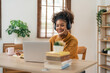 © Natee Meepian - Distance Education. Portrait of smiling woman african american sitting at desk, using laptop and writing in notebook, taking notes, watching tutorial, lecture or webinar, studying online at home