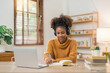 © Natee Meepian - Distance Education. Portrait of smiling woman african american sitting at desk, using laptop and writing in notebook, taking notes, watching tutorial, lecture or webinar, studying online at home