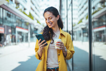 Modern Young Woman Walking On The City Street Texting And Holding Cup Of Coffee. Business Woman Holding Smartphone And Looking Away Outdoors. Beautiful Woman Spending Time In The City