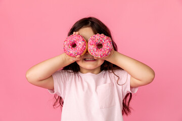 Portrait of a little smiling girl with curly hair and two appetizing donuts in her hands, closes her eyes with donuts, on a pink background, a place for text. Dieting concept and junk food.