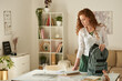 © Mediaphotos - Attractive redhead student girl standing at table in living room and packing textbook into satchel
