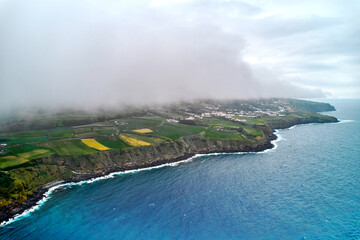 Wall Mural - Rocky shoreline, misty clouds over agricultural fields. Azores