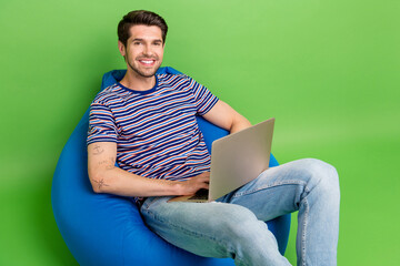 Poster - Photo of cute positive guy wear striped t-shirt sitting bean bag communicating modern gadget isolated green color background