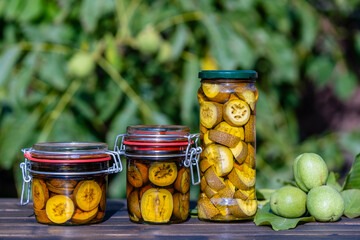 Wall Mural - Tincture of green walnuts in a glass jars on a wooden table in a summer garden. Sliced unripe walnuts in alcohol in a jar, to prepare homemade tincture, closeup