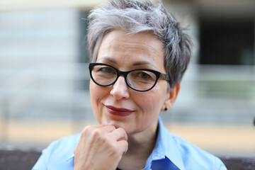 Sticker - A senior woman smiles with confidence. She is wearing a blue collared shirt and has gray hair. She is resting her hand on her chin and looking at the camera.