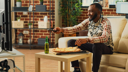 Wall Mural - African american guy enjoying pizza from delivery, binge watching favorite tv show in living room. Young happy man eating fast food from takeaway place, sitting at home with action film.