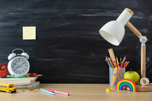 Learning Materials Layout. Side View Photo Of Pencils, Ruler, Stack Of Copybooks, Plasticine, Apples, Lamp And More On Desk Against A Chalkboard Background. Empty Space For Text Or Promotional Content
