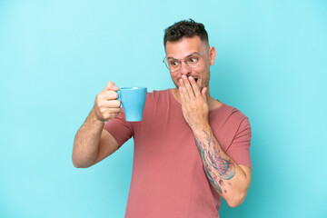 Wall Mural - Young caucasian man holding cup of coffee isolated on blue background with surprise and shocked facial expression