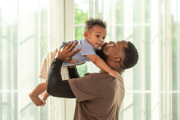 Wall Mural - Happy african black parents dad father throw baby son on piggyback and neck riding teasing fun near window. Black baby son and daddy enjoy teasing kiss cheek and neck riding at window light