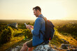 © baranq - Adult man using phone hiking during sunset sitting on top of the mountain