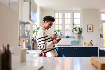 Mature Woman At Home In Kitchen Drinking Coffee And Checking Social Media On Mobile Phone