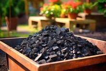 A Container Filled With Biochar Stands In A Garden With Green Plants And Colorful Flowers In The Background. Biochar Increases Soil Carbon To Improve Agricultural Productivity And Soil Fertility