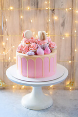 Tender and elegant pink cake decorated with melted white chocolate, cupcakes, meringues on festive background with string lights and bokeh