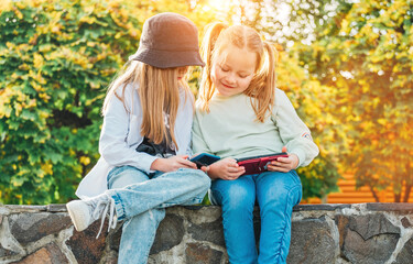 Two smiling little sister girls kids sitting and browsing their smartphone devices in the autumnal park. Careless young childhood time and a modern technology concept image.