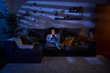 © CarlosBarquero - Young woman with happy expression looking at cell phone sitting in living room at night. Smiling cheerful latina female using mobile on sofa at home. Copy space, generation z and free time indoors.