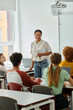 © LIGHTFIELD STUDIOS - Young smiling african american teacher with notebook talking to pupils during lesson in class