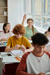 © LIGHTFIELD STUDIOS - Teenage schoolgirl raising hand and talking near devices and classmates during lesson in school