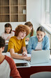 © LIGHTFIELD STUDIOS - Smiling teenage classmates using laptop together near notebooks during lesson in classroom in school