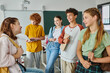 © LIGHTFIELD STUDIOS - Positive teenage pupils with devices and backpacks talking while standing in classroom in school