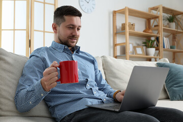 Poster - Happy man with cup of drink working with laptop on sofa at home