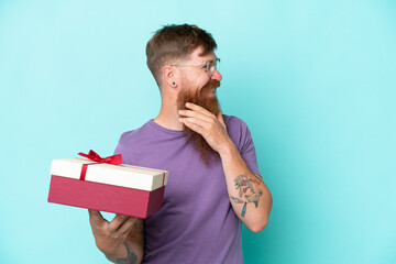 Wall Mural - Redhead man with long beard holding a gift isolated on blue background thinking an idea and looking side