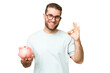 © luismolinero - Young man holding a piggybank over isolated chroma key background showing ok sign with fingers