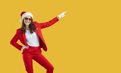 Joyful cheerful confident pretty young woman in stylish red suit, Christmas cap and trendy sunglasses standing isolated on yellow background, pointing to copy space side, inviting to party and smiling