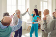 Friendly nurse, caregiver or psychologist woman giving high five to senior man during therapy in session with group of elderly people sitting in circle. Psychological support in nursing home.