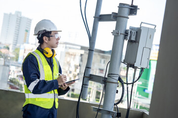 Building inspector man using digital tablet while checking telecommunication pole or telecom tower. Asian male technician worker inspecting cellular phone network antenna on rooftop of the building