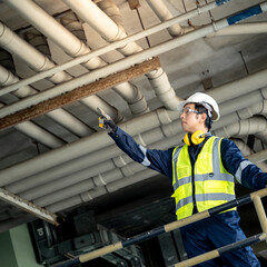 Asian male building inspection engineer pointing at plumber piping system. Construction worker man with reflective vest, safety helmet, protective goggles and ear muffs working at at construction site