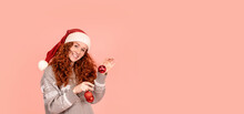 Cheerful Christmas Young Woman In Christmas Sweater And Santa Hat Holding Christmas Decoration. Merry Christmas And Happy New Year Concept