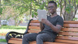 © stockbakers - Young African Man Doing Video Chat on Tablet while Sitting Outdoor on a Bench