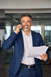 © Stock 4 You - Middle aged Latin or Indian businessman walking, having call on smartphone with business partner and holding documents. Mature Hispanic man talking by mobile cellphone at work in office, vertical