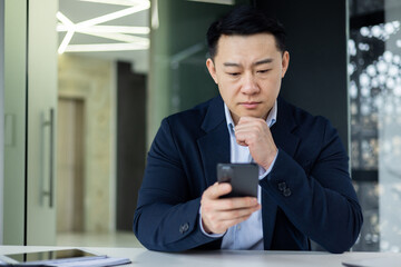 Wall Mural - Mature thinking asian boss inside office workplace with phone in hands, successful businessman thinking sitting with laptop in business clothes.