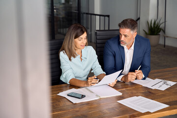 Wall Mural - Diverse partners mature Latin businessman and European businesswomen discussing project with documents at table in office. Team of colleagues professional business people working together, copy space