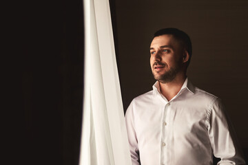 Wall Mural - Portrait of young man in white shirt in hotel room posing looking at window. Confident guy groom dressing getting ready to meet bride. Concept of young family, happy married. Copy ad text space