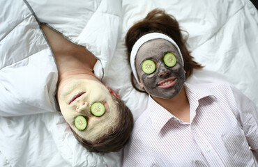 Wall Mural - Top-view of man and woman making spa procedures applying wetting facial masks and fresh cucumber on eyes. Couple laying on bed. Wellness and skincare concept